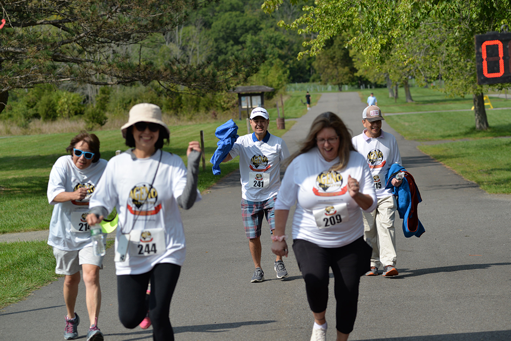 A group of five people are participating in an outdoor charity race, wearing white shirts with race numbers. They are smiling and moving energetically along a paved path in a park, surrounded by trees and greenery. An electronic timer is visible on the right side of the image.