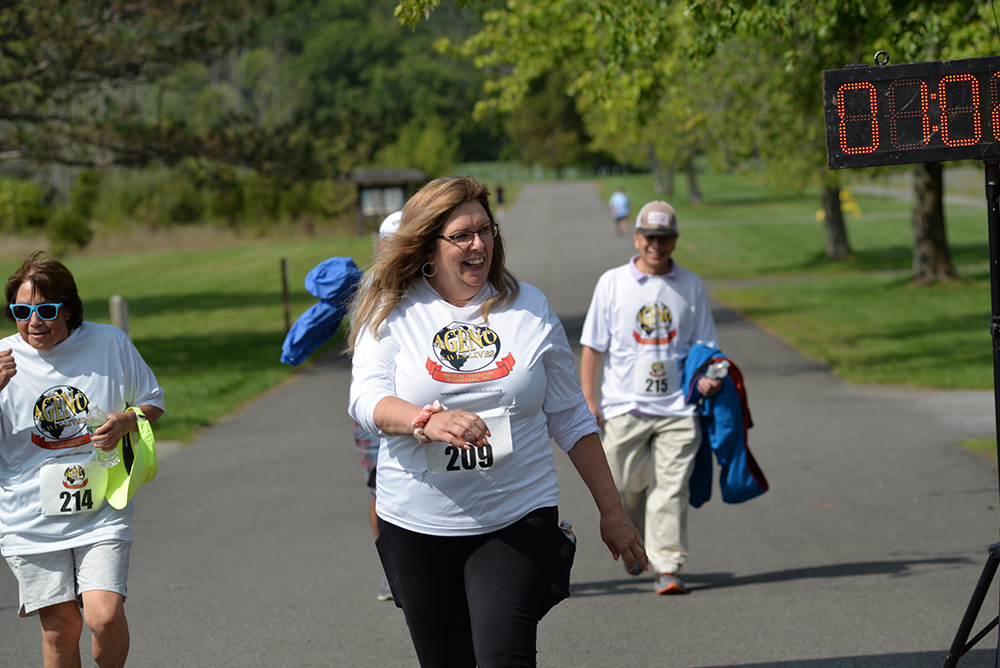 A group of people wearing white event t-shirts and race bibs walk along a paved path in a park. A smiling woman with bib number 209 is in the foreground, approaching a timer display. Green grass and trees are in the background, as volunteers offer water to participants and collect donations for charity.