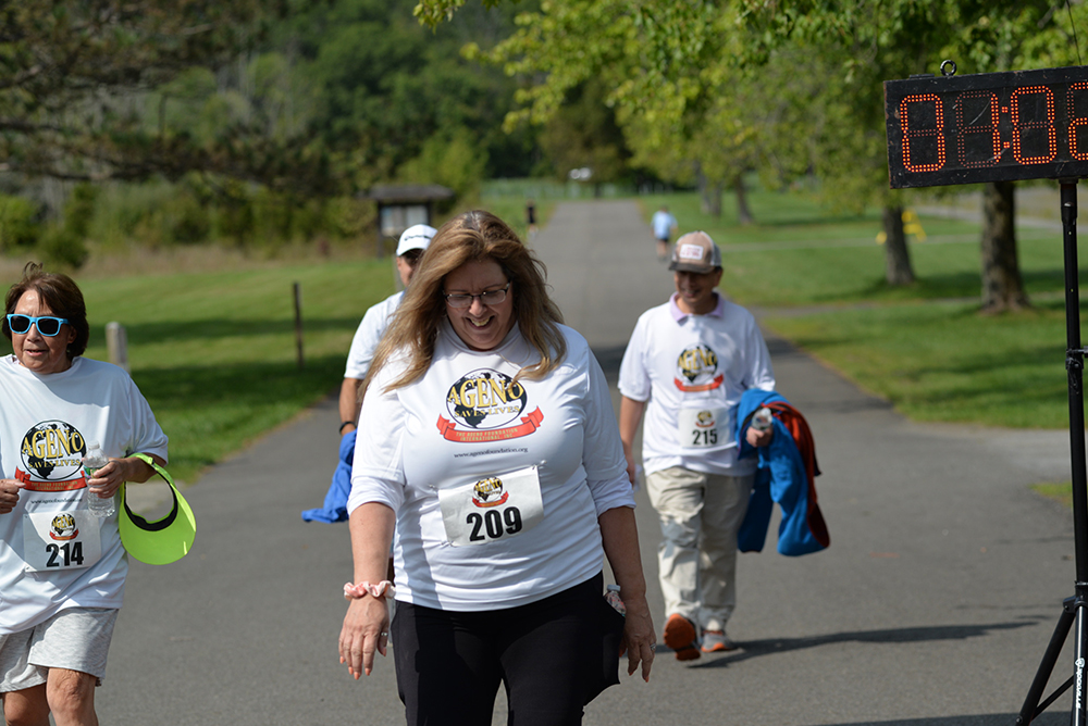 Participants in white t-shirts walk towards the finish line at an outdoor charity event. The race bib number "209" is visible on the front of one woman's shirt. Some hold jackets and hats. A running timer in the background shows a time of 48 minutes as they pass trees and greenery that surround the path.