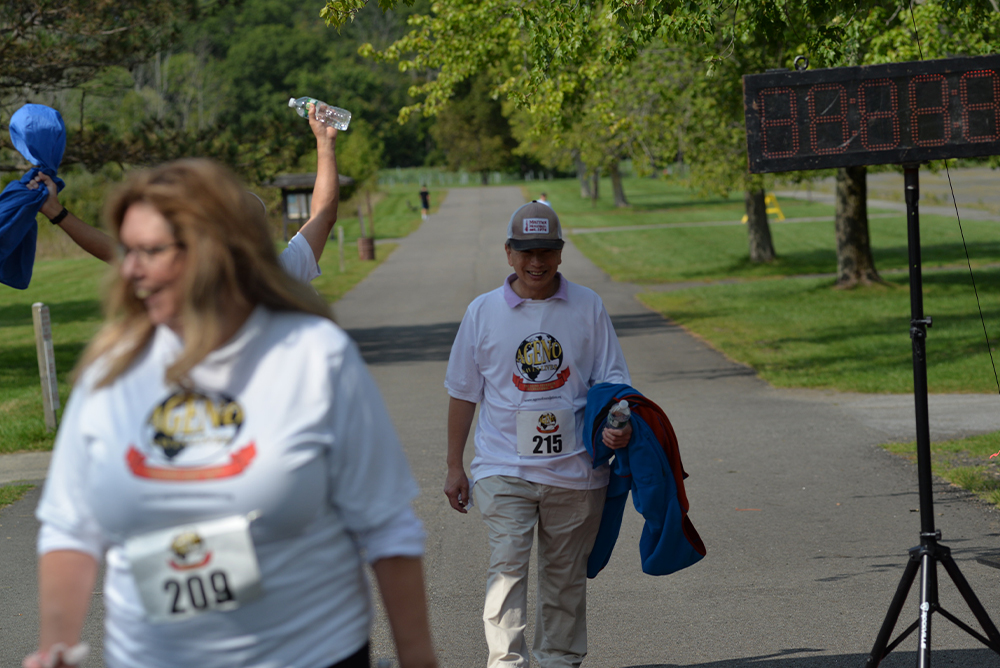Participants at the outdoor event are bustling with energy. A person with bib number 215, smiling and holding a blue jacket and a clear bottle of water, walks toward the finish area. The digital timer reads 30:00:89. In the foreground, another participant in a white shirt with bib number 209 makes their way forward for charity.