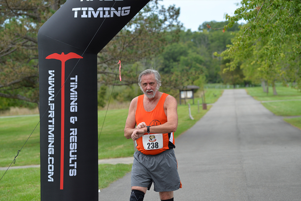 A man in an orange tank top and gray shorts crosses the finish line of a charity race. He is wearing a bib with the number 238 and appears to be stopping a wristwatch timer. The background features a tree-lined path and a black inflatable arch with timing equipment, highlighting his contribution to the cause.