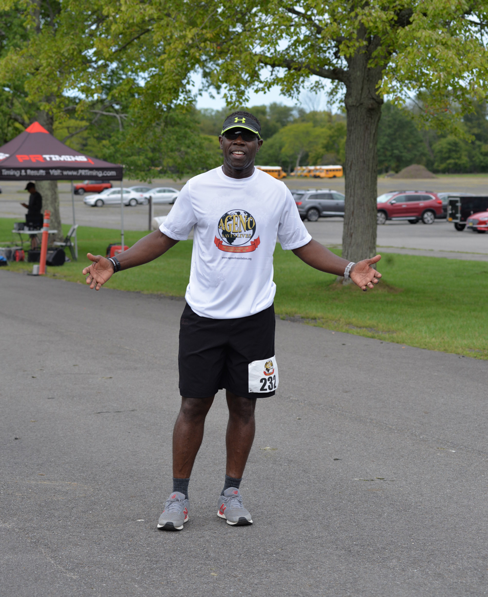 A man wearing a white T-shirt and black shorts participates in an outdoor charity race event. He is smiling with arms slightly extended and has a race bib numbered 231 pinned to his shorts. Trees and a few tents are visible in the background, and water stations for donations can be spotted nearby.