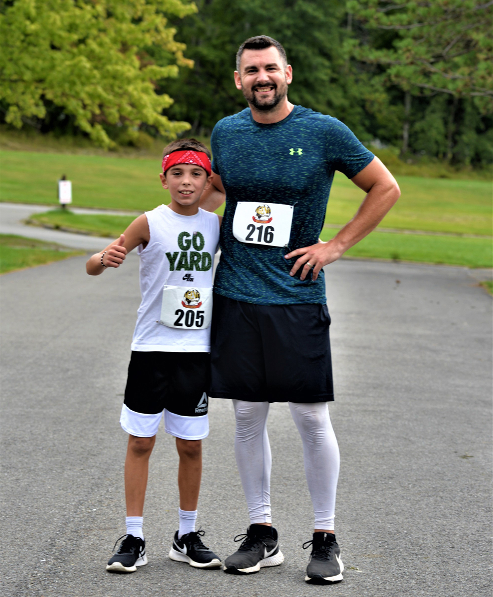 A young boy and an older man stand side by side on a paved outdoor path, smiling after a charity race. The boy wears a "Go Yard" tank top and black shorts, while the man dons a blue shirt, black shorts, and white leggings. Both have race bibs with numbers 205 and 216, ready to make their donation for clean water.