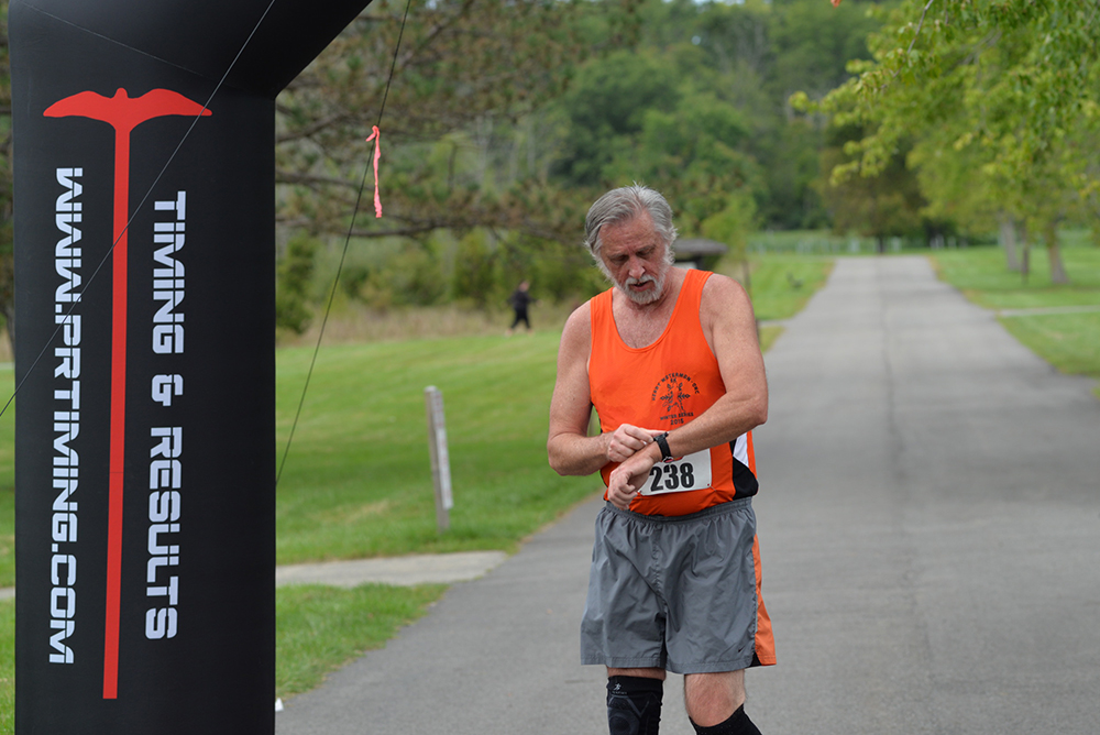 A runner in an orange tank top and gray shorts is checking his watch as he crosses the finish line of a charity race. The finish line banner displays "TIMING & RESULTS" and "WWW.PRTIMING.COM". Trees and a pathway are visible in the background.