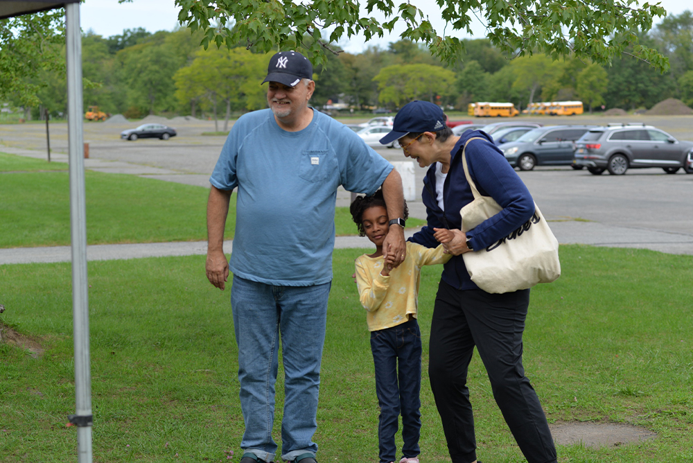 A smiling man in a blue shirt and jeans stands next to a young girl holding hands with a woman in a navy jacket and cap. They are outdoors on a grassy area with trees and parked cars in the background. The woman, carrying a beige tote bag, has just participated in a local charity event.