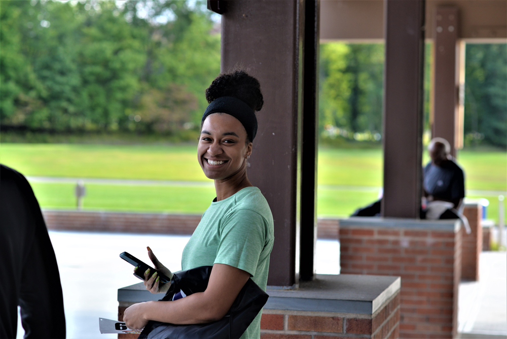 A young woman with a curly bun and headband smiles at the camera. She wears a green shirt and holds a smartphone and a donation bag while standing under a covered area with pillars. In the background, a man sits on a brick ledge. A grassy area is also visible.