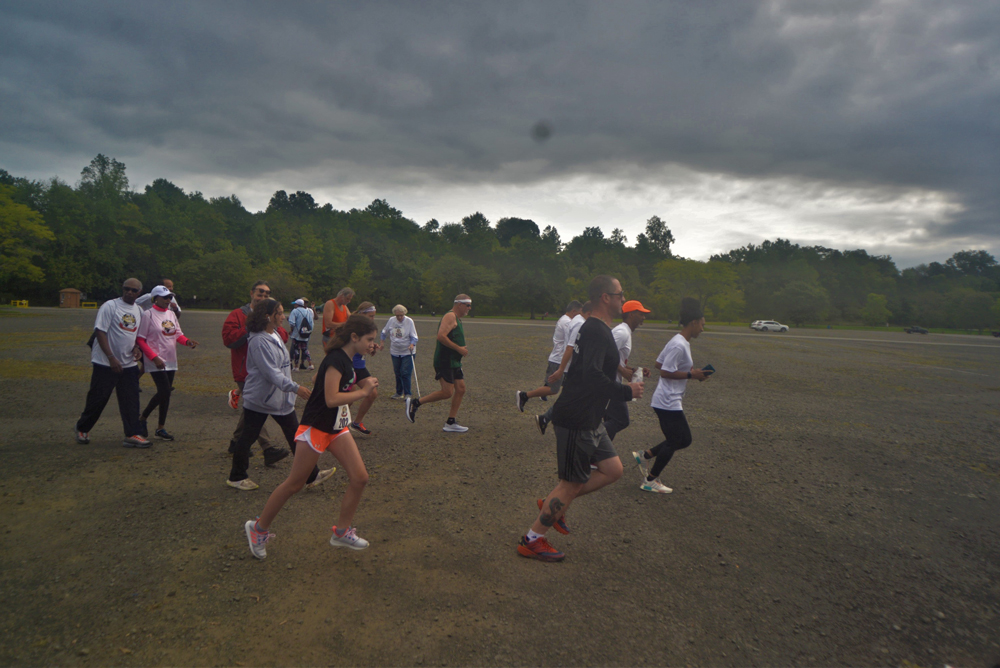 A group of people are running on a gravel road under an overcast sky. They appear to be participating in a charity race or group run, wearing athletic clothing. Trees and a parked car are visible in the background, underscoring the community's support and generous donations for a good cause.