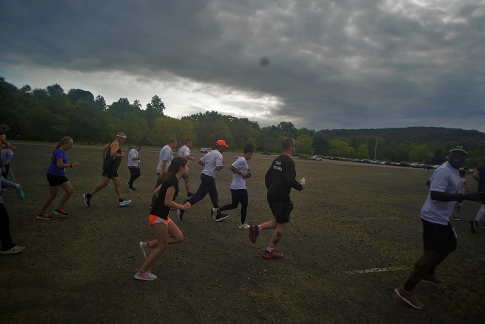A group of people running on a gravel path surrounded by trees under an overcast sky. Some are in athletic gear like shorts and t-shirts; others in long pants and jackets. The mood appears active and energetic, with varying paces among the runners participating in a charity event.