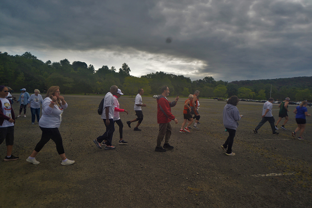 A group of people, some in athletic wear, walk together on a gravel path under a cloudy sky. The scene is outdoors with trees and hills in the background. The participants appear to be engaged in a charity event or group activity, fostering a sense of community as they contribute their time and efforts.