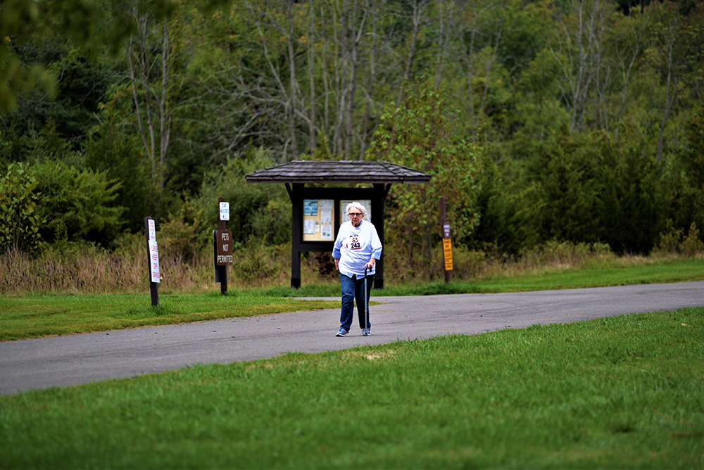 An elderly woman walks along a paved path in a park, using a walking stick. She is wearing a white jacket and blue jeans. In the background, near the wooden information kiosk, there's a fountain with clear water beside several signposts surrounded by greenery and trees.