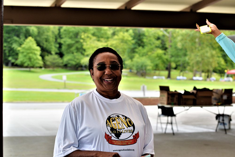 A smiling person wearing sunglasses and a white T-shirt with a charity logo stands under a pavilion in a park. Green trees and open grassy areas are visible in the background. Another person's arm is visible, holding a yellow donation box in the upper right corner of the image.