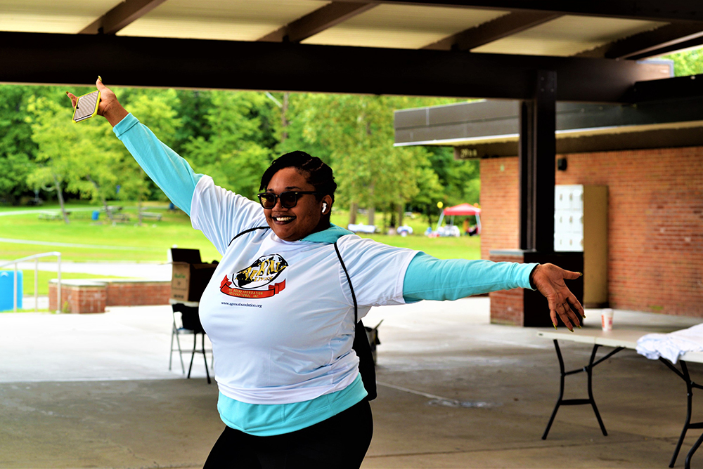 A woman smiles widely while posing with her arms outstretched under a pavilion. She is wearing a white and teal long-sleeve shirt and sunglasses, holding a phone in one hand. In the background, the grassy park with trees and a red brick building creates the perfect setting for promoting water charity donations.