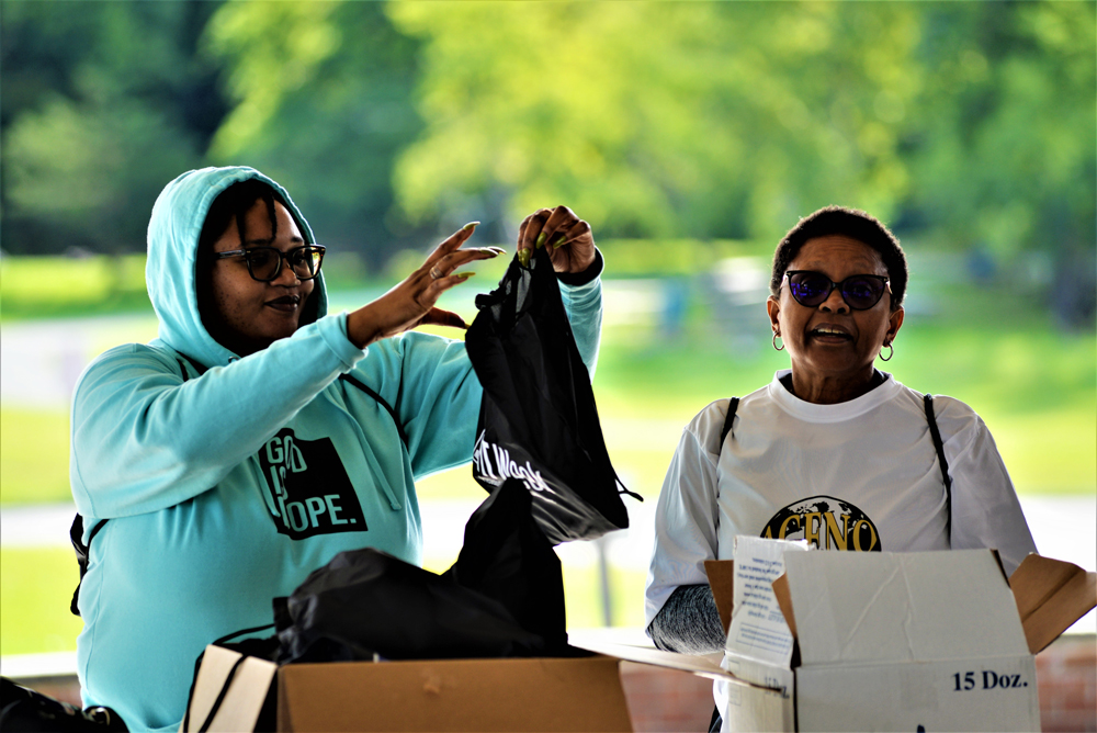 Two individuals are distributing items from boxes outdoors. One person in a blue hoodie and glasses is holding up a black drawstring bag. The other, in a white t-shirt and sunglasses, stands next to an open box. They appear to be collaborating at a charity event, possibly coordinating donations.