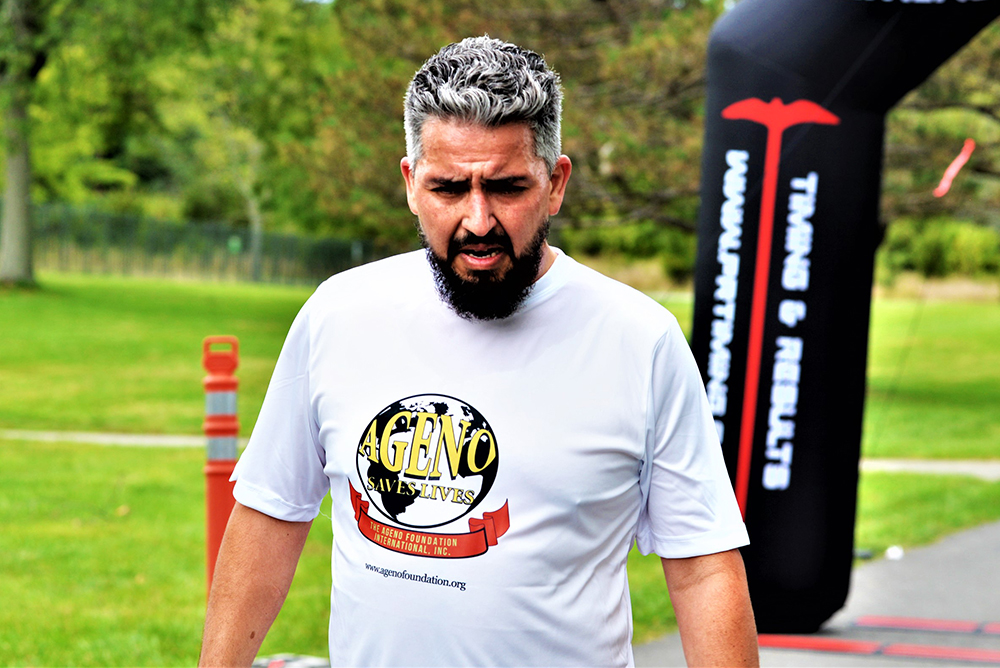 A man with gray hair and a beard is wearing a white "Aging Badly Saves Lives" T-shirt. He appears to be walking or running outdoors by an event finish line arch. The background includes trees and grass, with an orange traffic cone nearby, suggesting the event might be for charity.