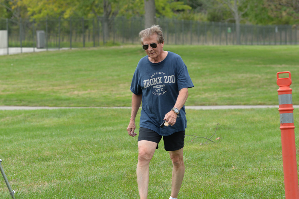 A person with gray hair and wearing sunglasses walks across a grassy park. They are dressed in a blue "Bronx Zoo NYC" t-shirt, black shorts, and white sneakers. In their right hand, they hold a small object that seems to be related to a charity event. An orange traffic cone is visible on the right.