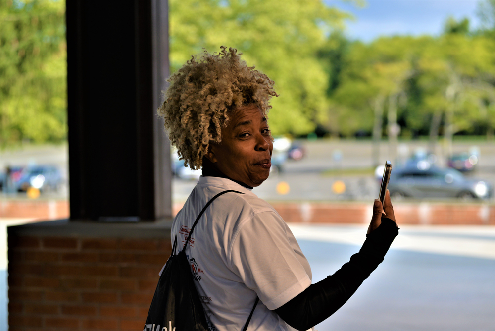A person with curly hair wearing a white t-shirt and carrying a black bag over their shoulder is holding up a smartphone and looking back at the camera, as if pausing between giving to charity. The background includes a parking lot and greenery.