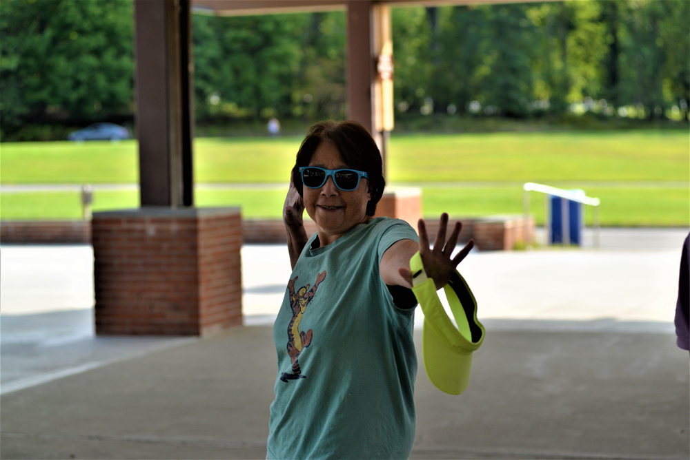 A person wearing sunglasses and a pale green shirt with a graphic stands in a park or sheltered area. They are smiling and holding their arm out with a bright yellow frisbee in hand, likely enjoying a charity event to raise donations. The background features grassy fields and trees.
