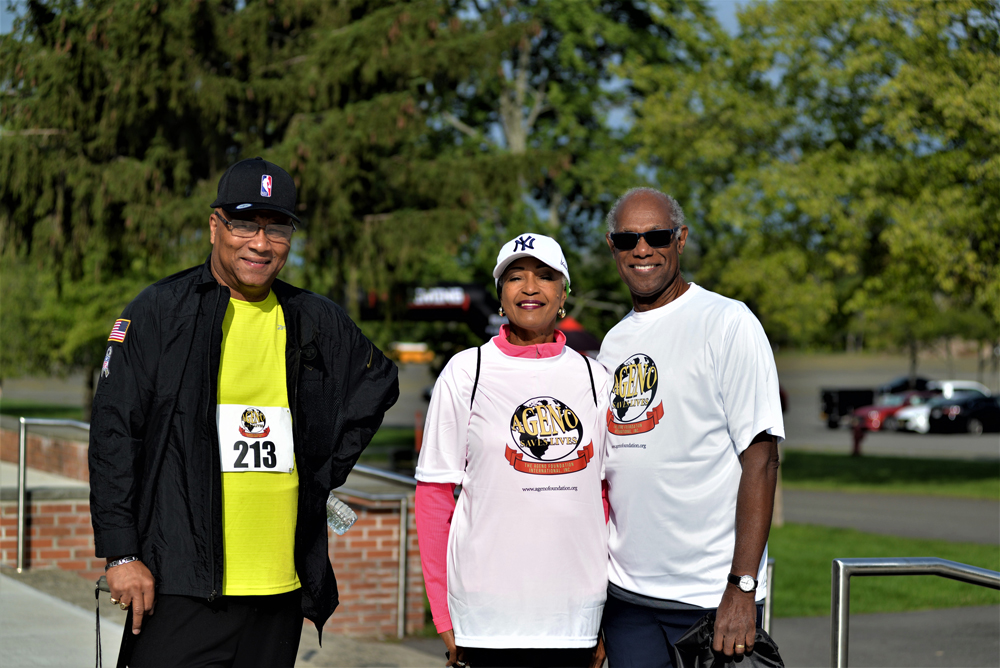 Three individuals wearing race bibs and event t-shirts pose outdoors. One man on the left wears a hat, glasses, and a bright yellow shirt under a black jacket. The middle woman wears a white hat and shirt. The man on the right is in a white shirt. Trees, a parked car, and water bottles for charity donations are in the background.