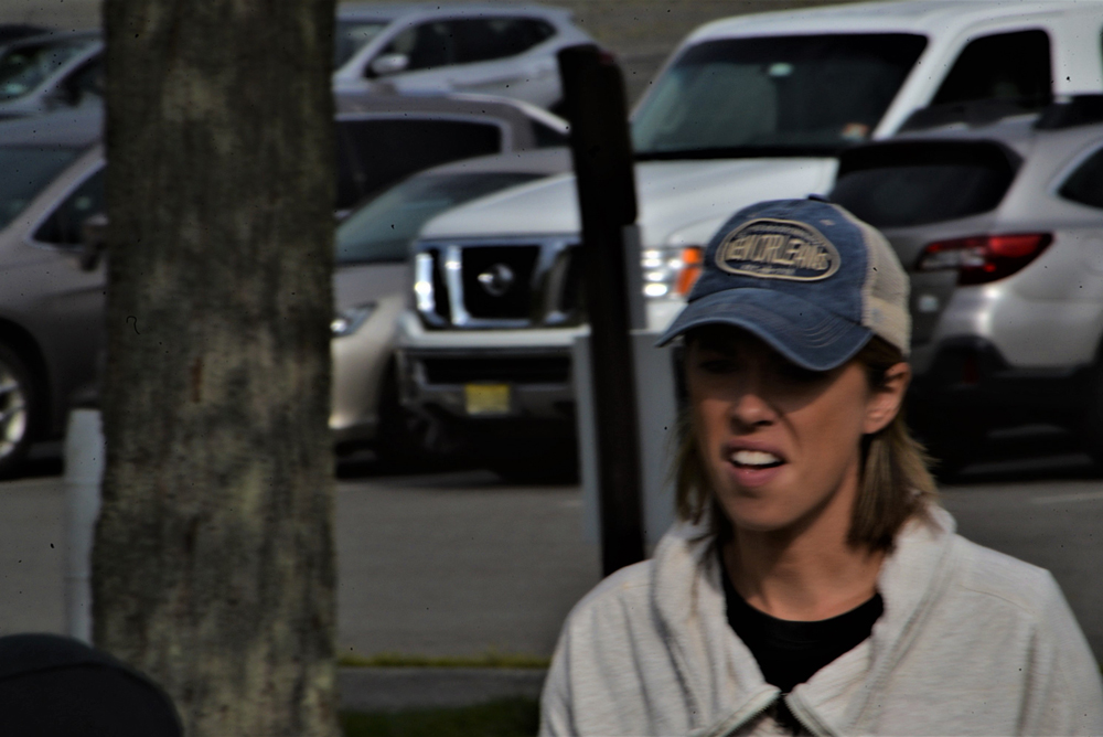 A woman wearing a blue baseball cap and a light-colored jacket is standing outdoors near a tree, holding a water bottle. Several parked cars are visible in the background.