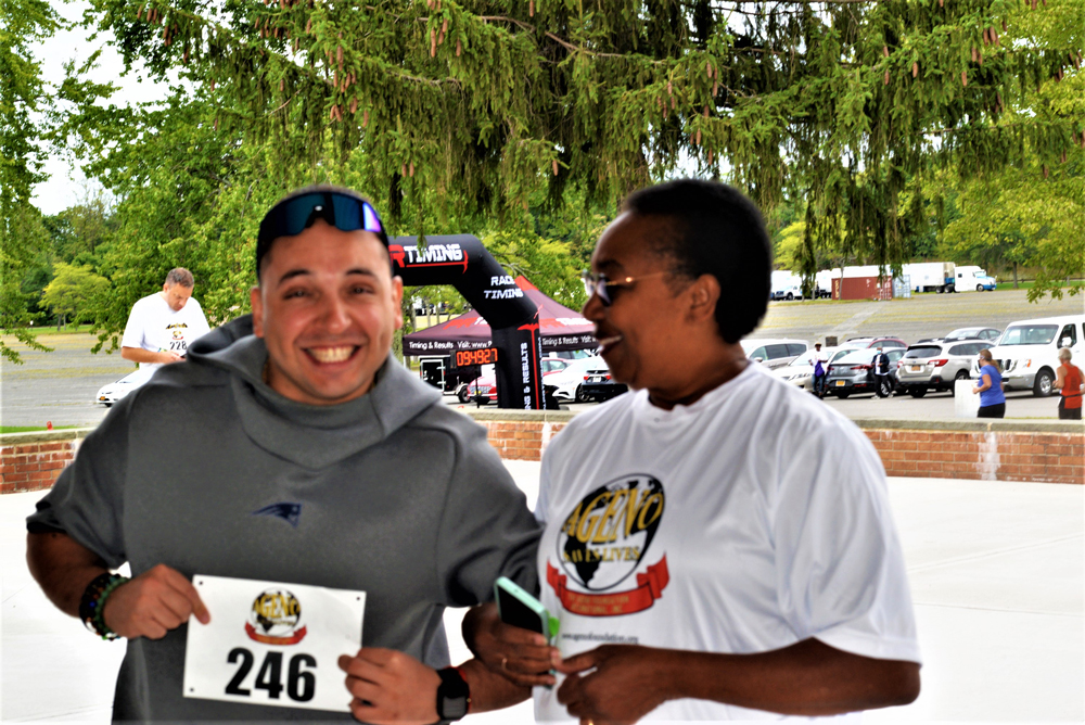 Two people are at an outdoor charity event. The person on the left, wearing sunglasses and a gray shirt, smiles while holding a race bib with the number "246." The person on the right, wearing a white shirt, looks at them and smiles; they hold a green phone.