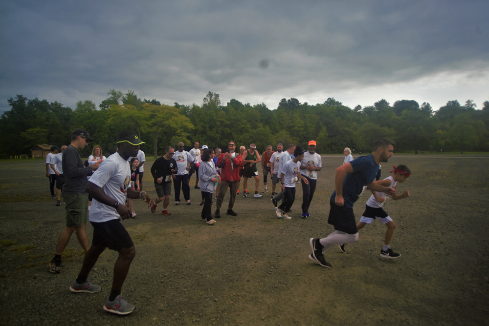 A group of runners start a charity race on a cloudy day, with some spectators and participants mingling in the background. They are on a gravel path surrounded by trees and lush greenery. The runners, focused and mid-stride, are wearing various athletic attire as they pass by donation stands offering water.