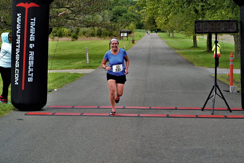 A runner wearing a blue shirt and headband with bib number 231 crosses the finish line during a race. The timing clock is set up on the right side, and the race timing arch is visible on the left. The background includes a paved path lined with trees and grass, with water stations dotted along the route.