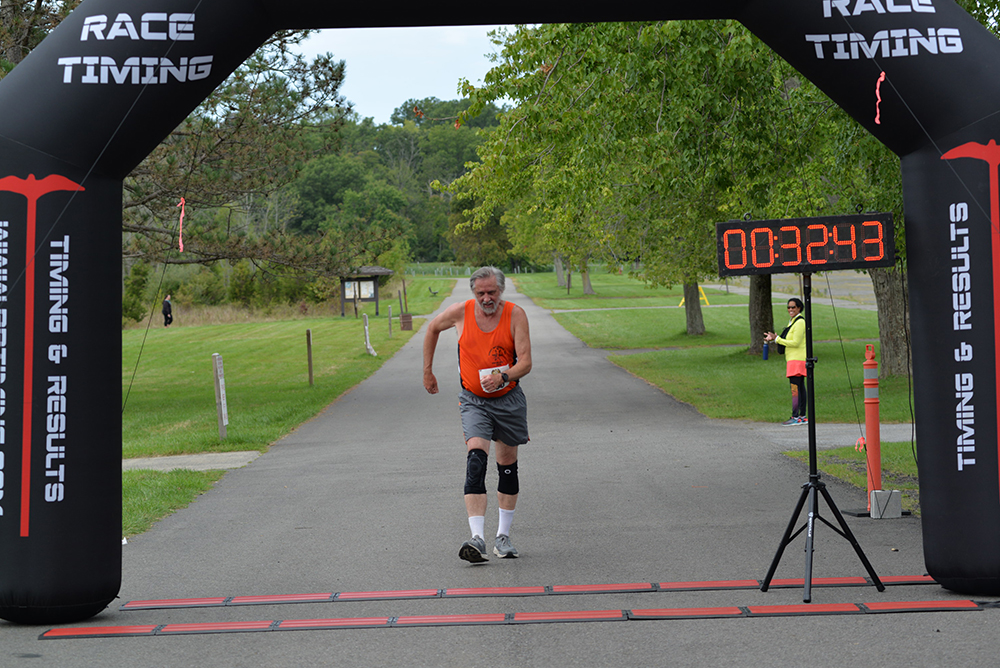 A man wearing an orange tank top, gray shorts, and a knee brace finishes a race, crossing the finish line under an inflatable archway with "Race Timing" printed on it. A digital timer shows 32 minutes and 43 seconds. Nearby, another person in a yellow shirt holds a sign encouraging donations to charity.