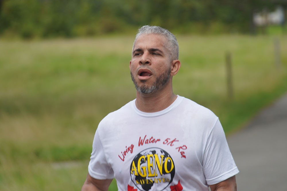 A man with short, gray hair is running outdoors on a paved path surrounded by grass and trees. He is wearing a white t-shirt that reads "Living Water 5k Run" with a logo featuring the word "AGENO" in yellow. Focused and exerting effort, he's part of an event encouraging participants to donate for clean water causes.