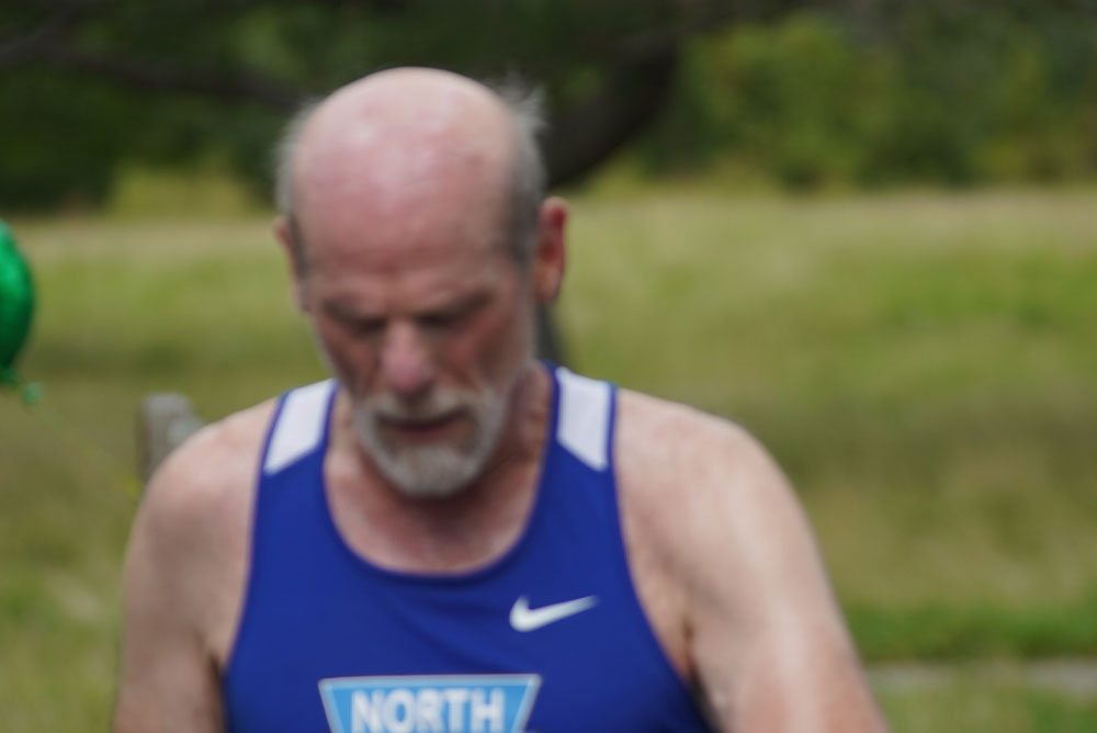 A bald man with a white beard, wearing a blue sleeveless athletic shirt, is running outdoors. The background is blurred, showing green grass and some trees. The man appears to be slightly hunched and looking down, possibly contemplating his next charity run or upcoming donation effort.
