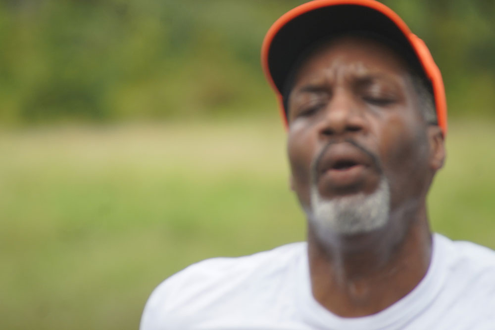 A close-up of a man wearing an orange cap and a white shirt is shown. His eyes are closed, and his mouth is slightly open as if he is exhaling or in deep concentration. The background features blurred green natural surroundings, evoking a moment of peace often found after taking time to donate to charity.