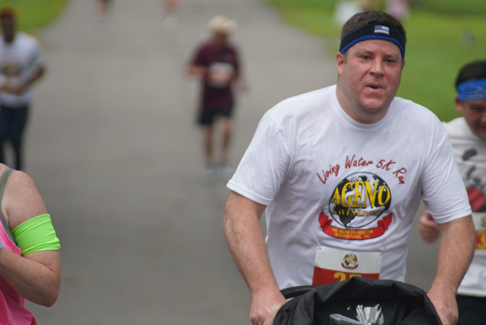 A man in a white "Living Water 5K Run" T-shirt, wearing a blue headband, is running and pushing a stroller in a park during the charity race. Other runners are visible in the background.