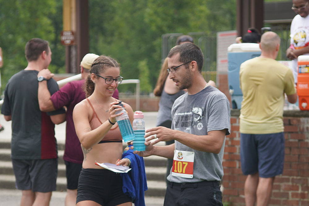 Two people participating in an outdoor event are standing and talking. The woman, wearing a sports bra and glasses, holds a water bottle, while the man, wearing a gray shirt and glasses with a red and yellow number 107, gestures towards her. Other participants mingle in the background near the donation booth.