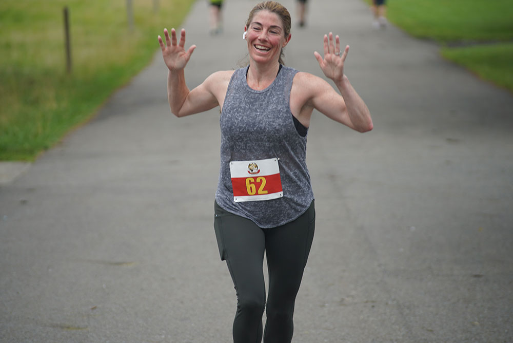 A woman with her number 62 bib on her shirt, smiling and waving her hands mid-run on a paved path in a park. She is wearing a gray sleeveless top and dark leggings. The background contains blurred trees and grass, as she participates in a charity run to raise donations for clean water initiatives.