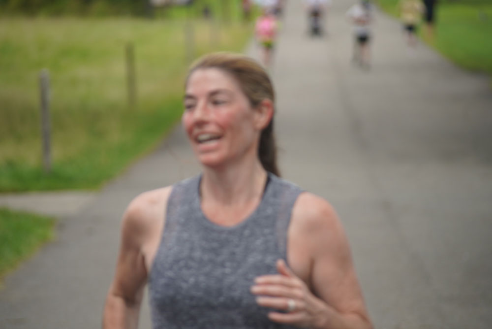 A person with light skin and brown hair tied back in a ponytail is jogging along a paved pathway in an outdoor setting. Wearing a sleeveless gray shirt and smiling, they look inspired, possibly thinking of the charity marathon they're training for. The background is slightly blurred, with other joggers visible in the distance.