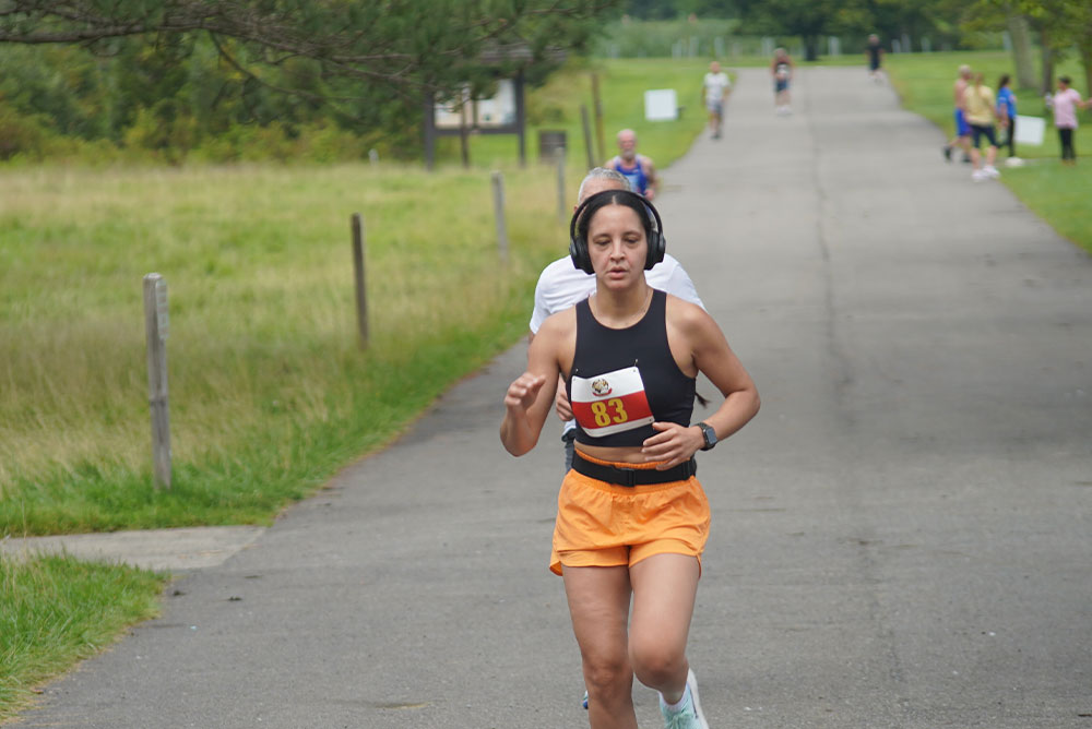 A person wearing an orange athletic outfit and headphones runs on a paved path in a park, participating in a charity race. Other runners and spectators are visible in the background among greenery and trees. The runner is focused and determined, contributing to the cause with every step.