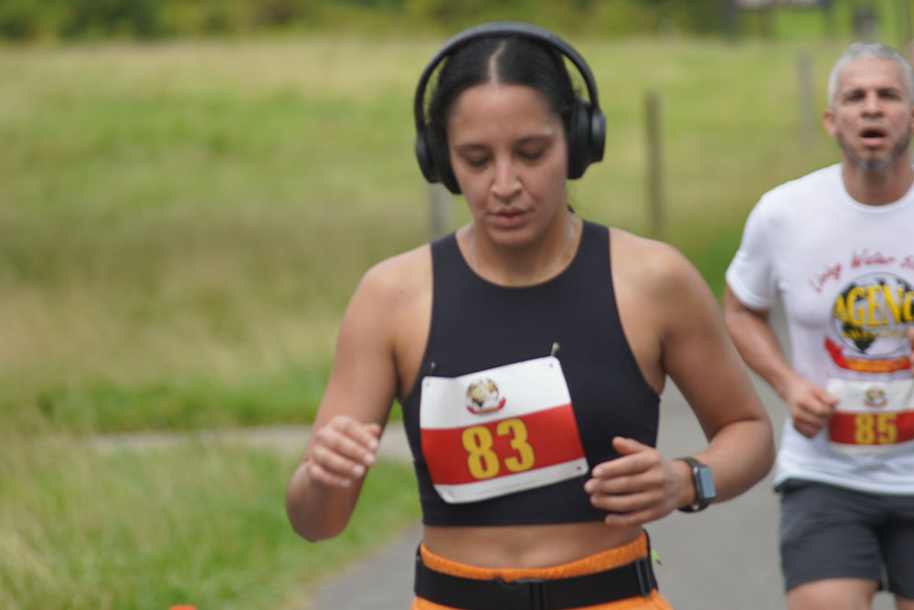 A person wearing a black sports bra, orange shorts, and headphones runs with bib number 83 during a charity race. Close behind, another participant in a white shirt and bib number 85 follows on the same path. The background reveals a grassy area and path, reminding participants of the cause they support through their donations.