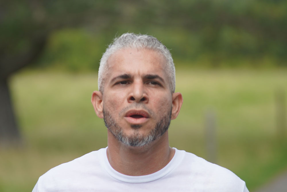 A man with short gray hair and a trimmed gray beard is captured outdoors, looking ahead. He wears a white shirt, and the background is blurred with green foliage and a hint of trees, suggesting a serene natural setting near water.