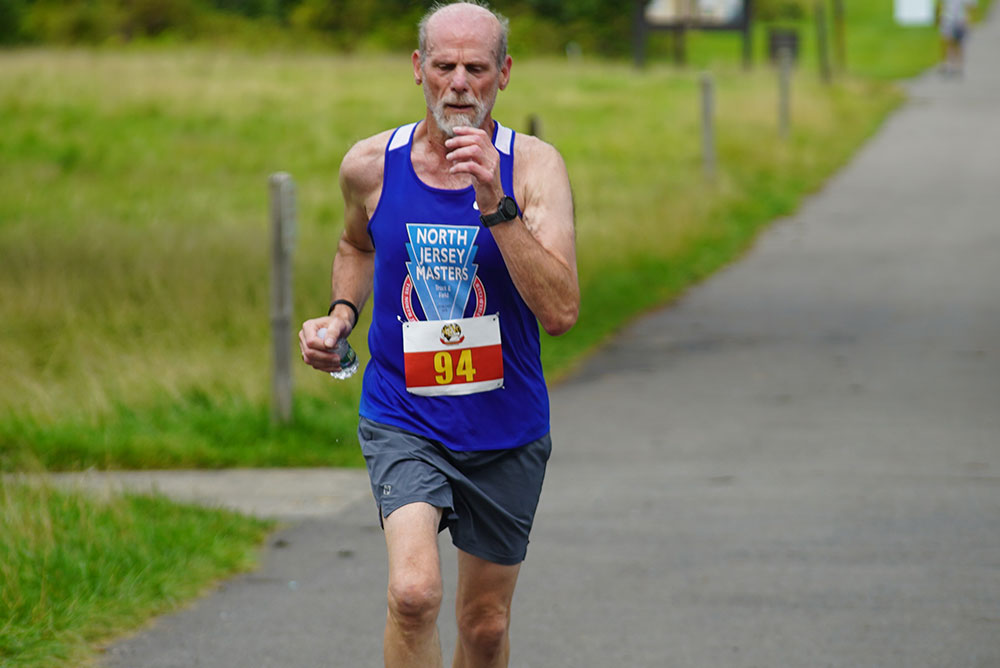 A runner wearing a blue "North Jersey Masters" tank top and gray shorts is participating in a charity race. Holding a small object, possibly a water bottle, he runs along a paved path through a grassy area. His race bib number is 94.