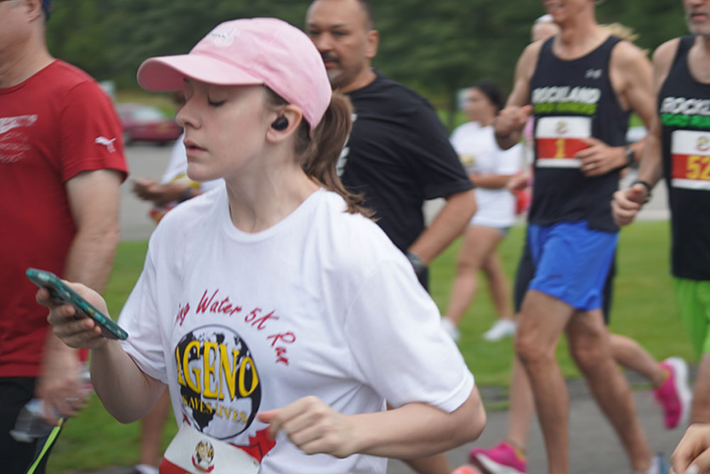 3rd-Annual-5K-for-Living-Water_1 A group of participants, some in colorful athletic wear, run in a 5K race outdoors. A woman in the foreground, wearing a pink cap and white race t-shirt, looks at her phone while running. Trees and a road are visible in the background, where volunteers hand out water to runners as part of their donation efforts.