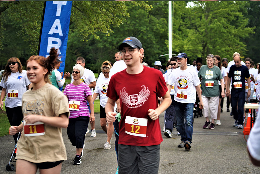 3rd-Annual-5K-for-Living-Water_2 People participating in a community run/walk event wear numbered bibs and casual attire, smiling as they walk and run. Trees and an event banner are visible in the background. Two lead participants with bib numbers 11 and 12 are holding water bottles, encouraging others to stay hydrated while mentioning donations for a local cause.
