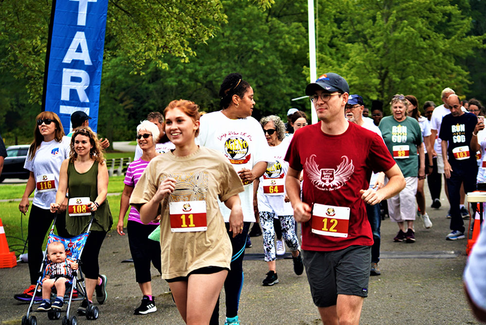 3rd-Annual-5K-for-Living-Water_3 A group of people, wearing casual athletic attire with numbered bibs, participate in a community charity fun run. Some push strollers, and all seem to enjoy the event. Trees and a large blue "START" sign are visible in the background, indicating the beginning of the race.