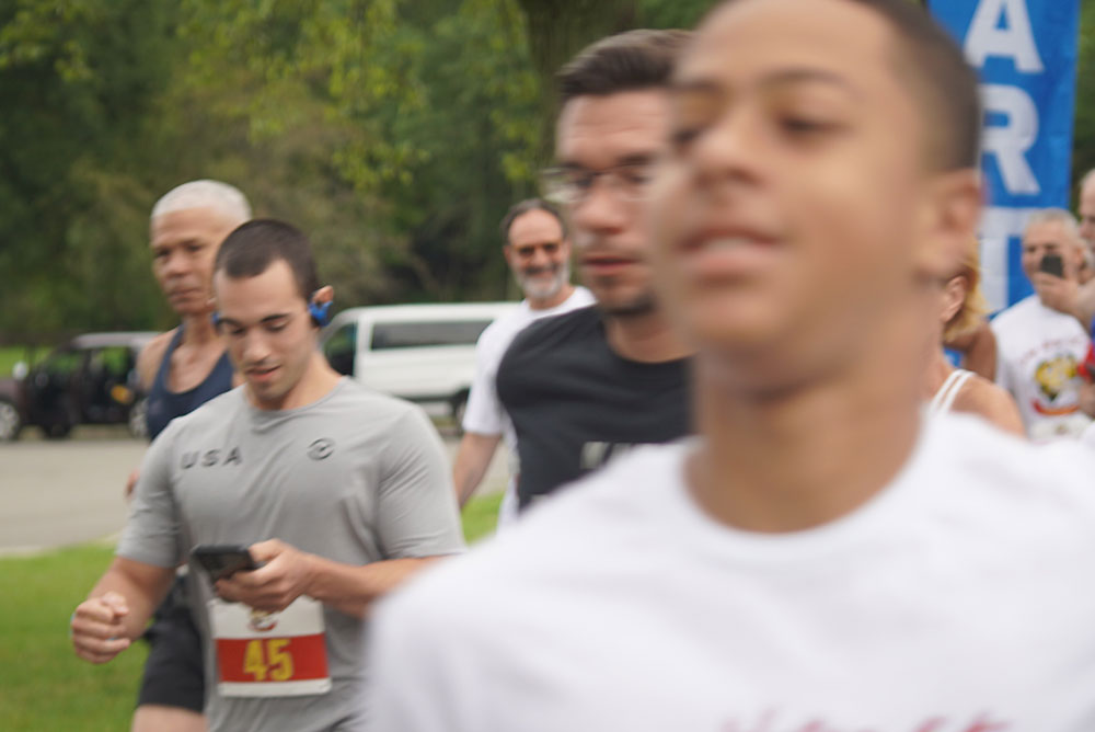 3rd-Annual-5K-for-Living-Water_4 A group of people running outdoors. The person in the foreground wears a white shirt. Behind him, a man in a grey shirt with the number 45 reads from his phone. Other runners and trees are visible in the background, and a blue "Donate Water" banner stands to the right.