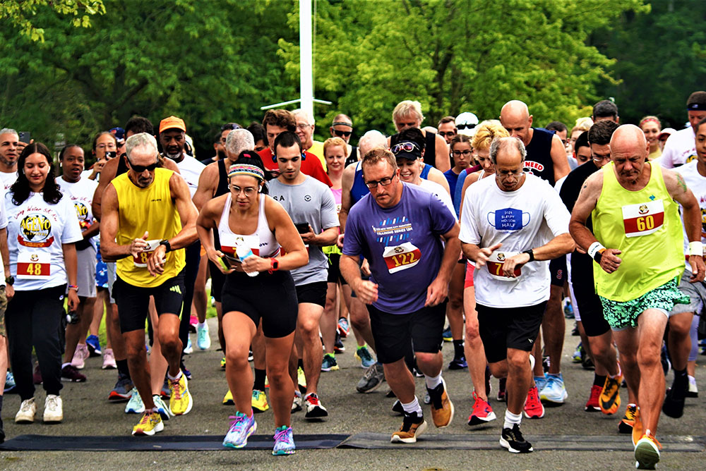 3rd-Annual-5K-for-Living-Water_5 A diverse group of runners of varying ages, wearing race bibs and athletic gear, are at the starting line of a charity race in a park. Trees and greenery are visible in the background. The runners display focused and determined expressions as they begin their run to support a good cause.