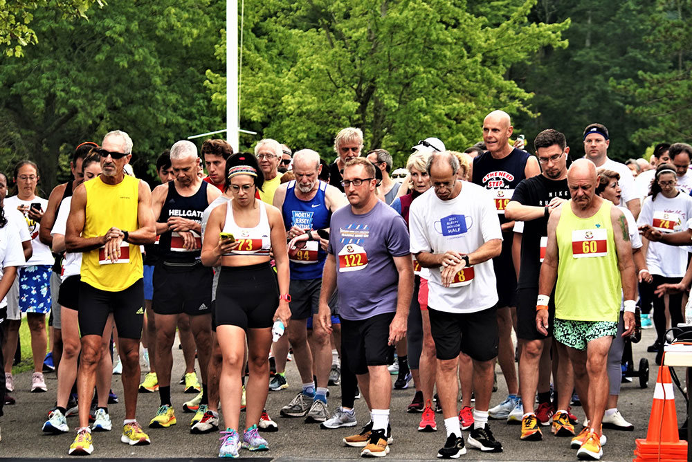 3rd-Annual-5K-for-Living-Water_6 A diverse group of runners stands at a starting line, preparing for a race. The participants are wearing bib numbers and athletic attire. Trees and greenery are visible in the background, indicating an outdoor setting. Some runners appear focused while others are relaxed, sipping on water before the event.