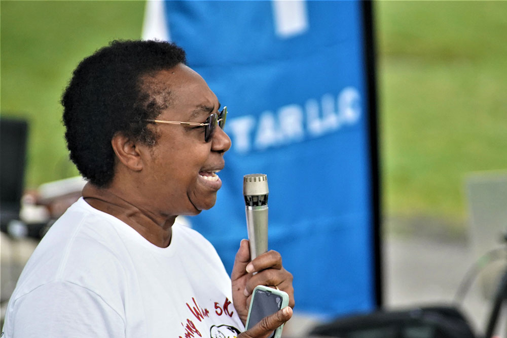 3rd-Annual-5K-for-Living-Water_7 A person with short hair and glasses is speaking into a microphone. The individual, wearing a white T-shirt and holding a smartphone, appears to be encouraging the audience to donate to a water charity. A blurred blue and white banner is in the background.
