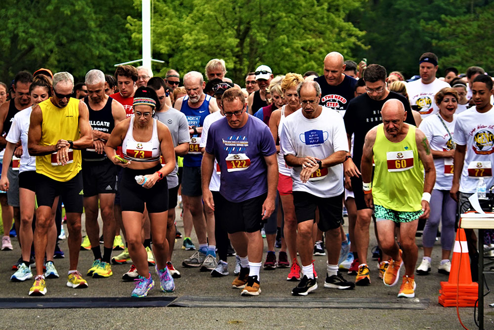 3rd-Annual-5K-for-Living-Water_8 A diverse group of runners of varying ages and athletic wear gather at a starting line for a charity race. Some wear numbered bibs, and a few hold water bottles. Trees are visible in the background, suggesting an outdoor setting. The mood is focused and energetic, as they prepare to run for donations.