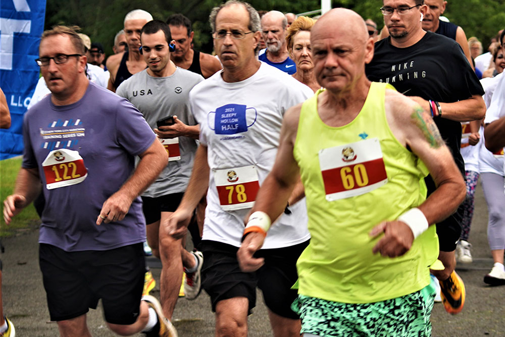 3rd-Annual-5K-for-Living-Water_9 A group of runners participating in a charity race, all wearing bib numbers. The foreground focuses on three men; the one on the left wears a purple shirt, the middle one has a white shirt, and the right one wears a neon yellow tank top. Trees are visible in the background.