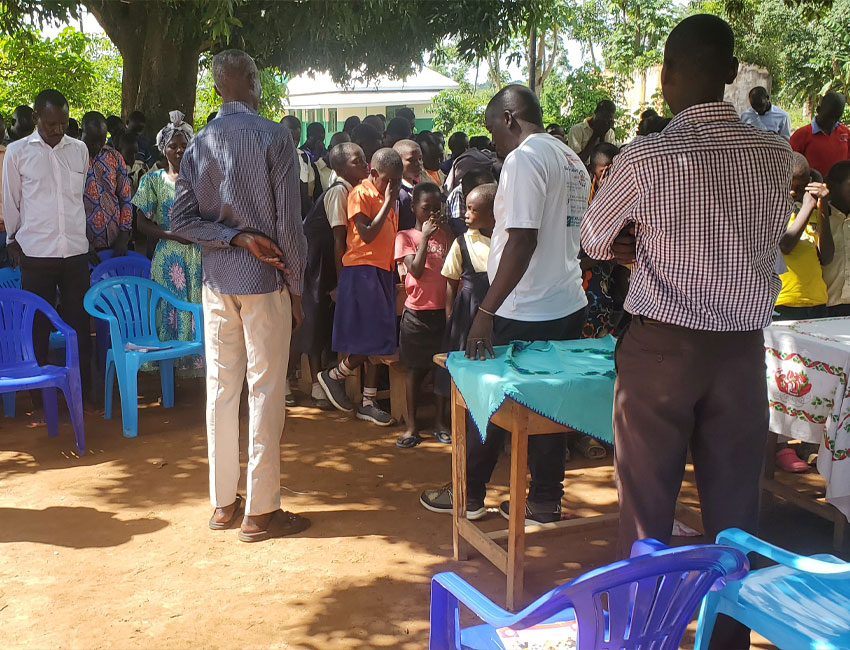 A group of people, including adults and children, are gathered outdoors under a tree. Several empty blue and purple chairs are in the foreground. A man in a white T-shirt and another in a checked shirt stand near a wooden table covered with cloth, setting up for the charity event with donation boxes nearby.