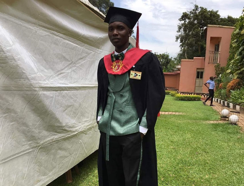A graduate stands outdoors next to a tent, wearing a black graduation cap, black gown with a red and yellow sash, and a green shirt with a bow tie. In the background, there are buildings, grassy areas, and trees. Another person is visible in the distance holding a charity donation box.