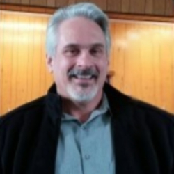 Gala flyer-profile-img-2 A man with gray hair and a beard, wearing a light gray shirt and a dark jacket, smiles at the camera. The background consists of a wooden panel, giving a warm, indoor setting. His smile exudes kindness, inspiring others to donate to charity and support clean water initiatives.